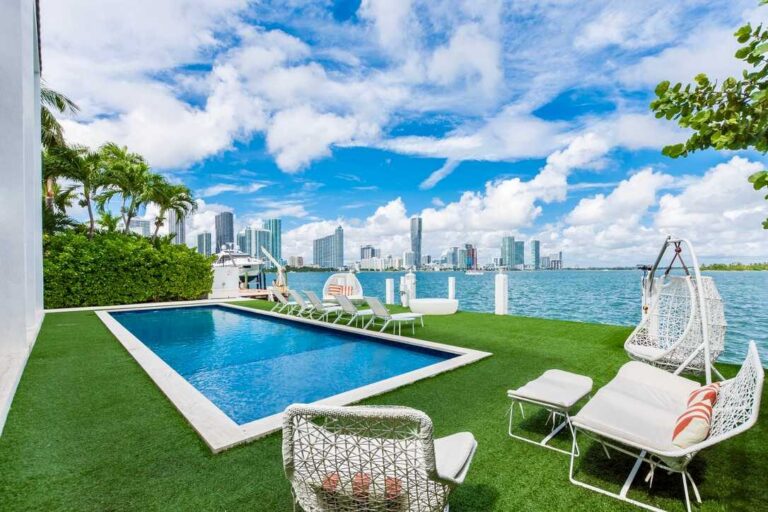 Waterfront outdoor space featuring a private swimming pool, artificial turf lawn, cushioned lounge seating, hanging egg chairs, poolside sun loungers, glass safety fencing, and panoramic bay and city skyline views from the terrace.