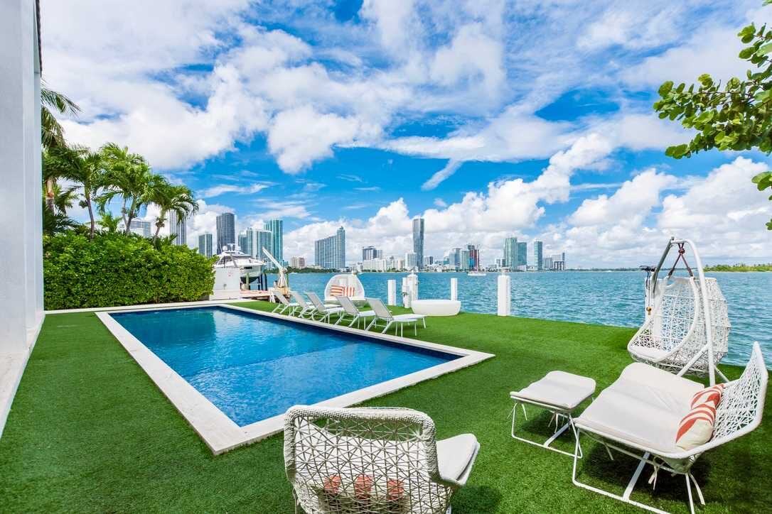 Waterfront outdoor space featuring a private swimming pool, artificial turf lawn, cushioned lounge seating, hanging egg chairs, poolside sun loungers, glass safety fencing, and panoramic bay and city skyline views from the terrace.