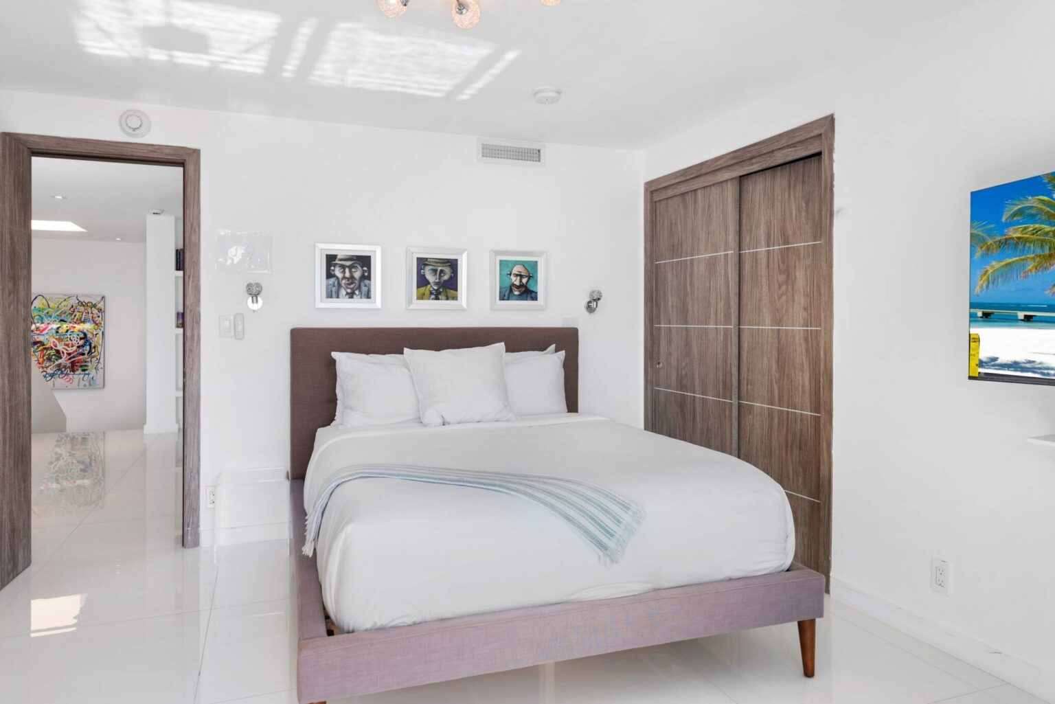 Comfortable guest bedroom featuring a queen-size bed with white linens, built-in closet storage, tile flooring, contemporary wall art, bedside reading lights, and a bright minimalist layout with direct access to the adjacent bathroom and living areas