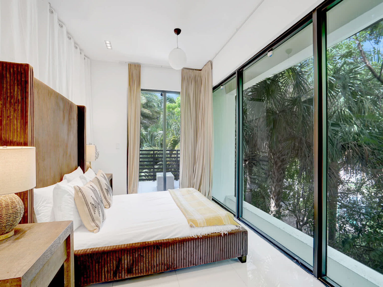 Spacious bedroom featuring a king-size bed, floor-to-ceiling windows, and sliding glass doors overlooking tropical greenery. Natural light, modern furnishings, and soft neutral finishes create a bright and comfortable retreat.