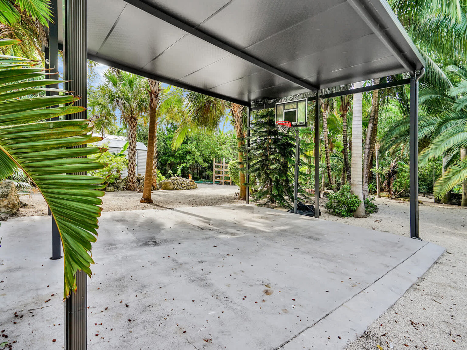 Covered parking area with a metal canopy, concrete driveway, and surrounding tropical landscaping. Positioned beside the home, this sheltered outdoor space provides convenient vehicle parking with easy access to the property.