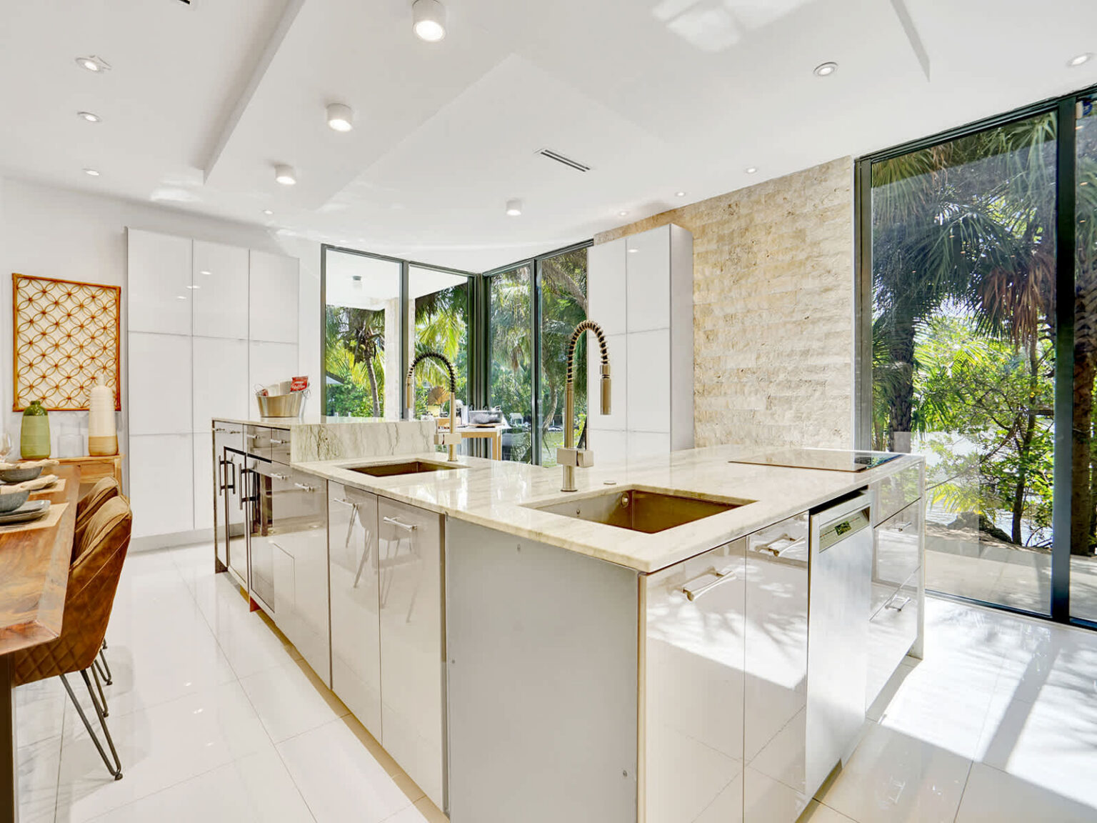 Bright modern kitchen featuring a large island with breakfast seating, sleek cabinetry, and polished countertops. Floor-to-ceiling windows provide natural light and tropical views, while the open layout connects seamlessly to the living and dining ar
