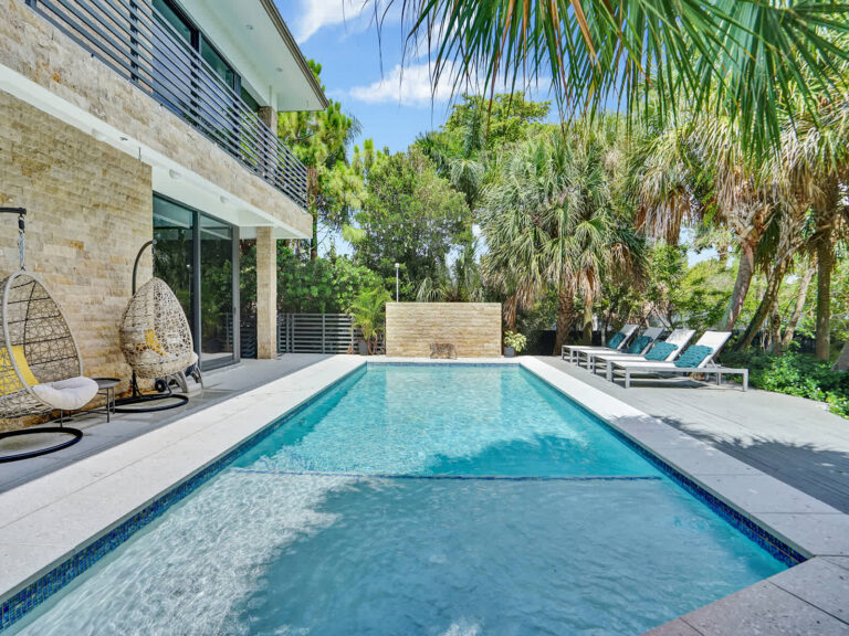 Rectangular swimming pool with a shallow entry area, spacious tiled deck, and direct access to the villa. Surrounded by tropical landscaping, mature palm trees, and lounge seating, this outdoor space offers peaceful garden views.