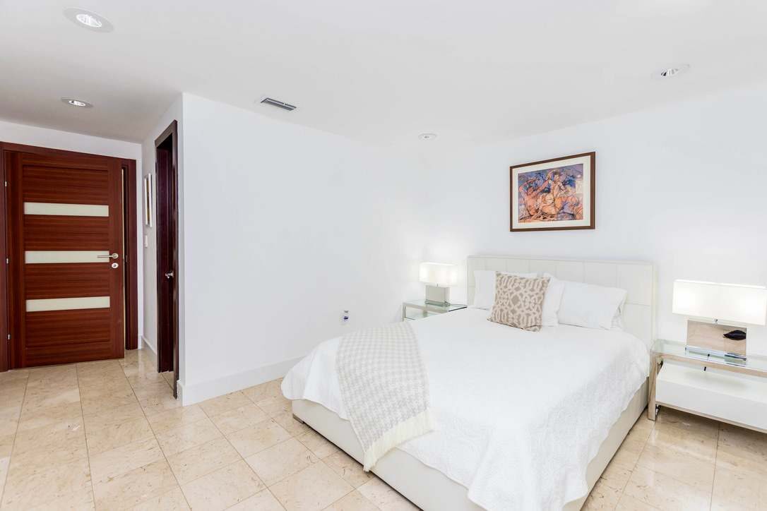Comfortable guest bedroom featuring a queen-size bed with white linens, bedside tables with lamps, tile flooring, recessed lighting, contemporary artwork, and a bright minimalist layout with convenient access to the adjacent bathroom and living areas
