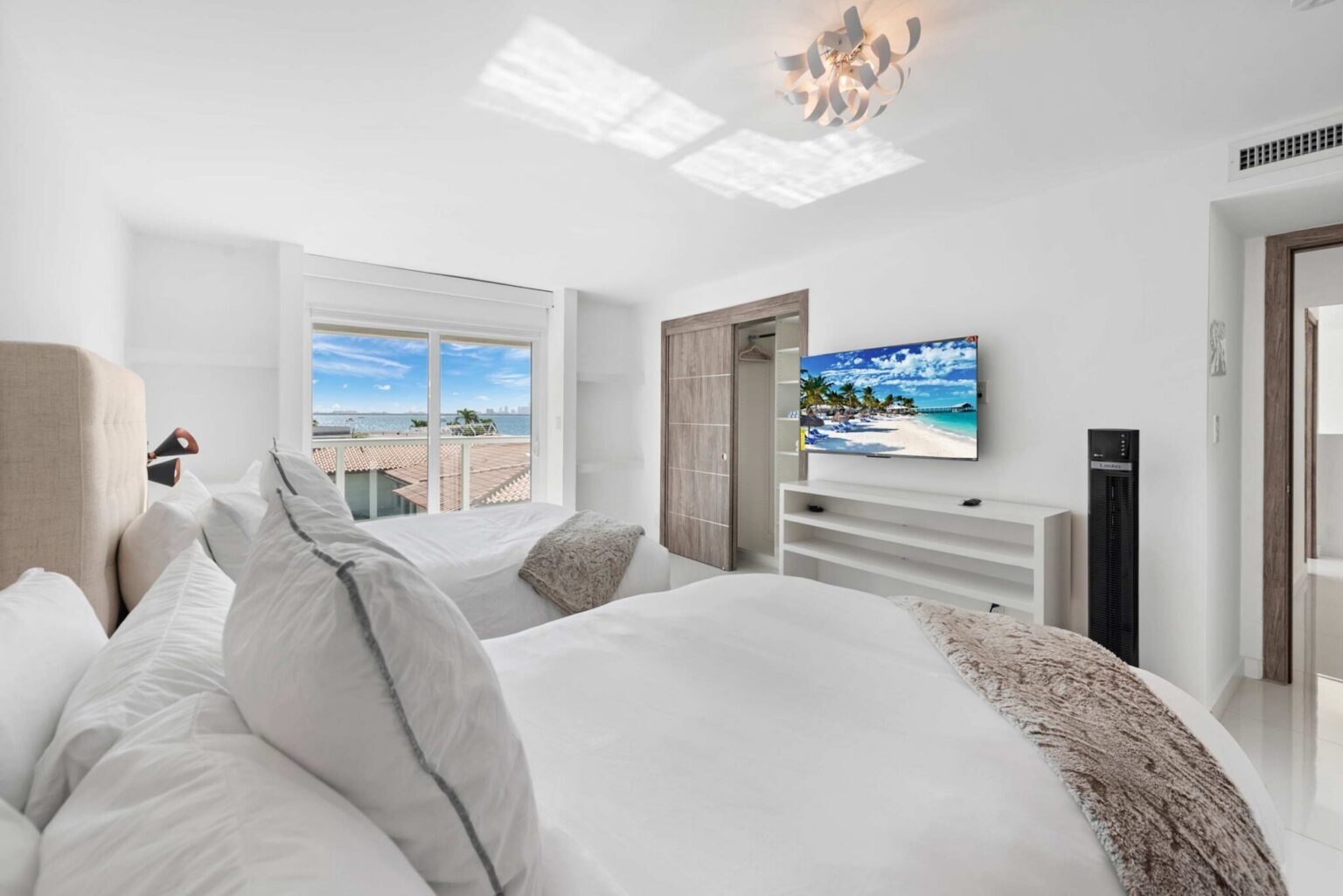 Waterfront guest bedroom featuring a queen-size bed with premium linens, wall-mounted smart TV, dresser storage, contemporary lighting, and a large picture window showcasing panoramic bay and city skyline views with abundant natural light.