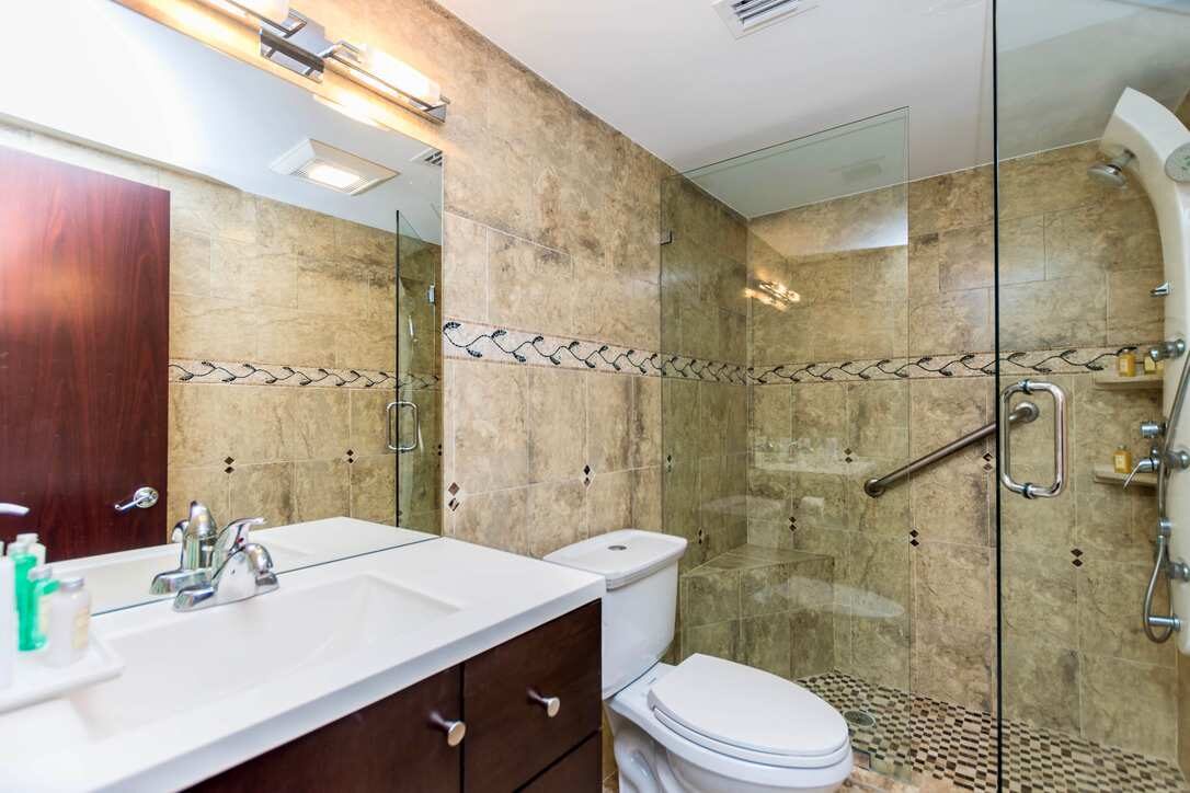 Full bathroom suite featuring a glass-enclosed walk-in shower, natural stone tile walls, oversized vanity with integrated sink, large wall mirror, modern lighting, built-in shelving, and contemporary fixtures designed for comfort and functionality.