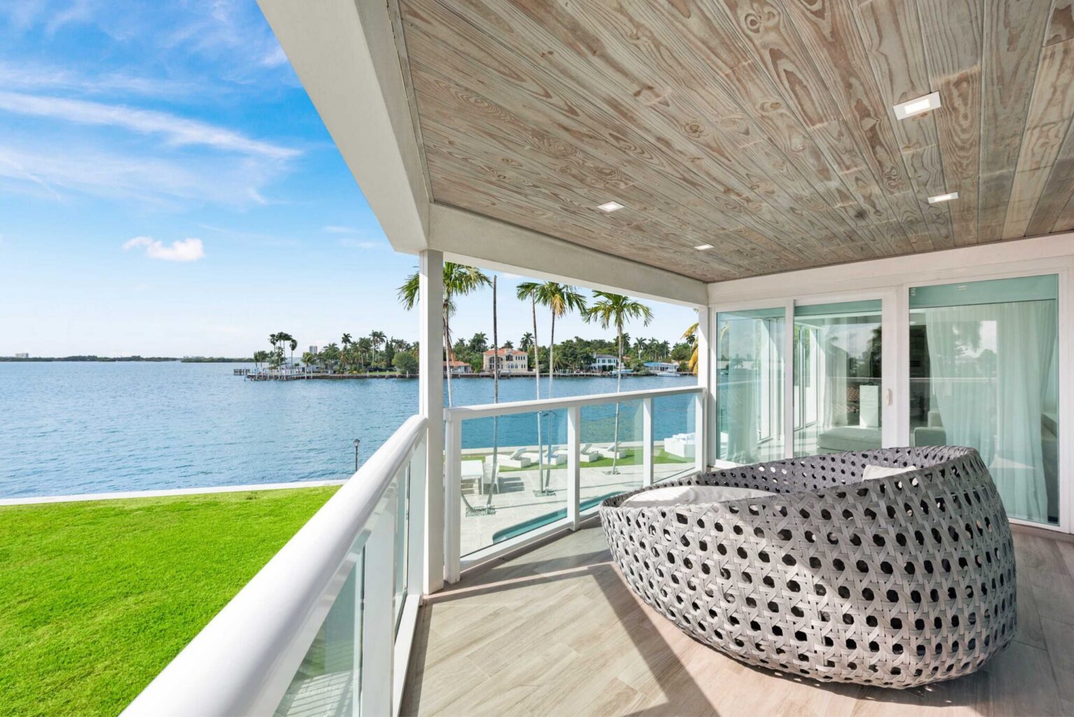 Covered waterfront balcony featuring woven outdoor lounge seating, glass railings, wood-look ceiling finishes, and panoramic bay views. Private outdoor space offers direct access from the bedroom and overlooks the shoreline and surrounding waterfront