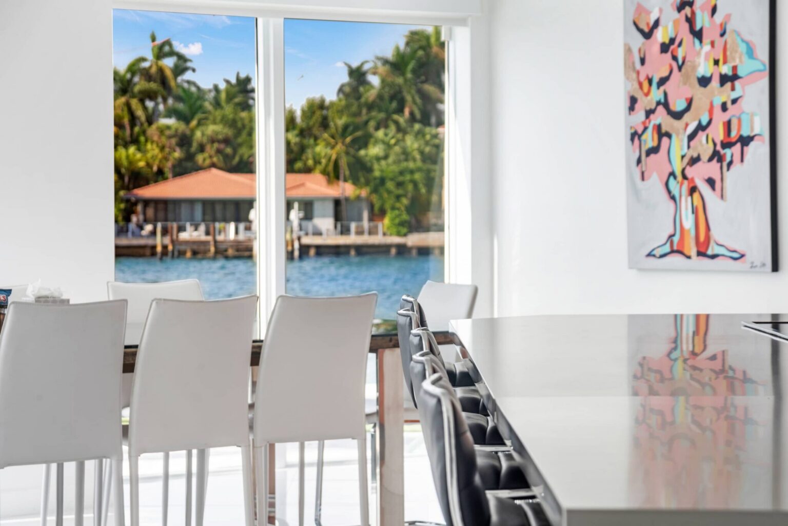 Dining area with seating for eight, waterfront views through large picture windows, white modern dining chairs, breakfast bar seating, contemporary artwork, and an open-concept layout connecting the kitchen and living spaces.