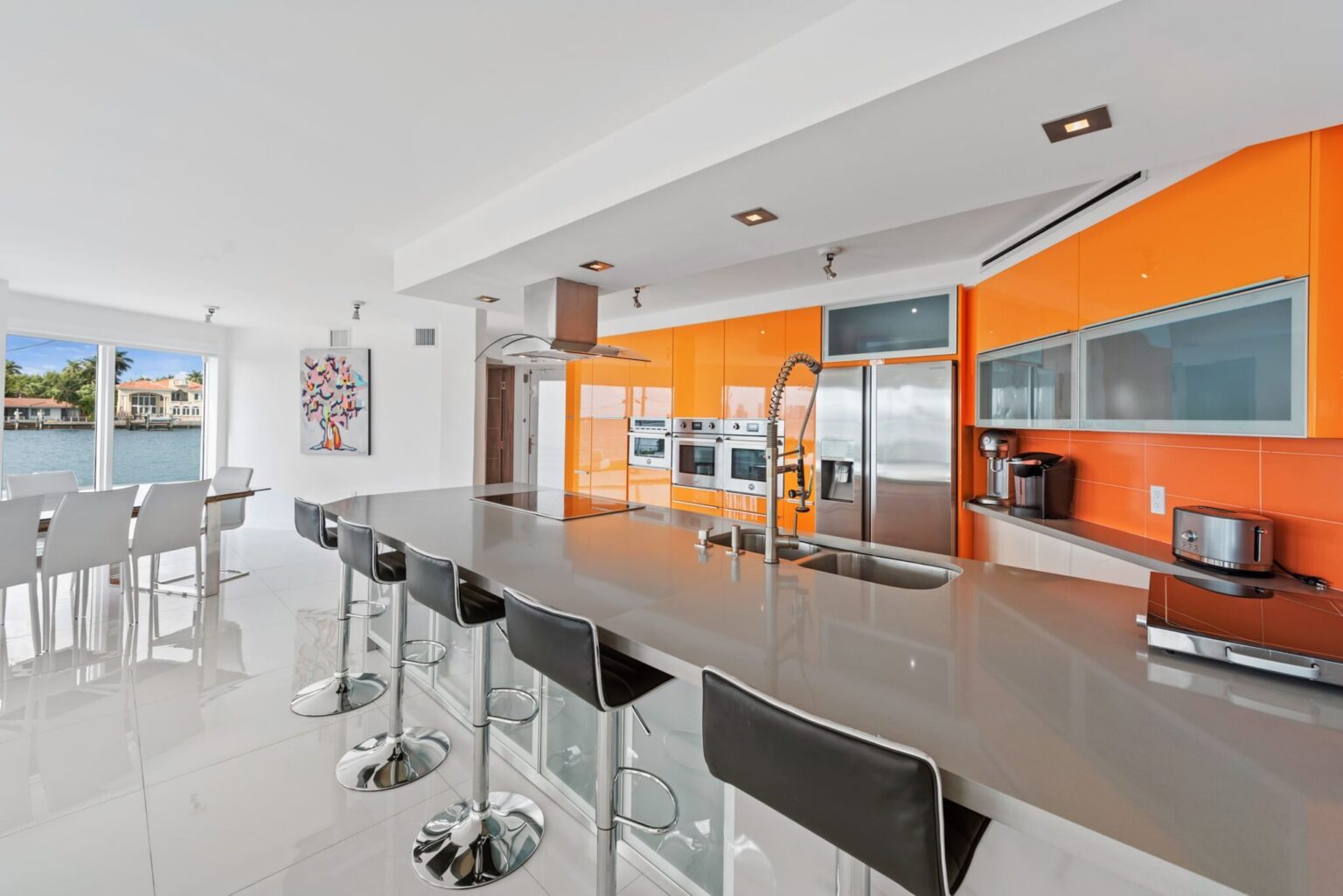 Chef-inspired kitchen featuring a large breakfast bar with seating, stainless steel appliances, custom high-gloss cabinetry, built-in wall ovens, expansive counter space, and an open-concept layout overlooking the dining and waterfront living areas.
