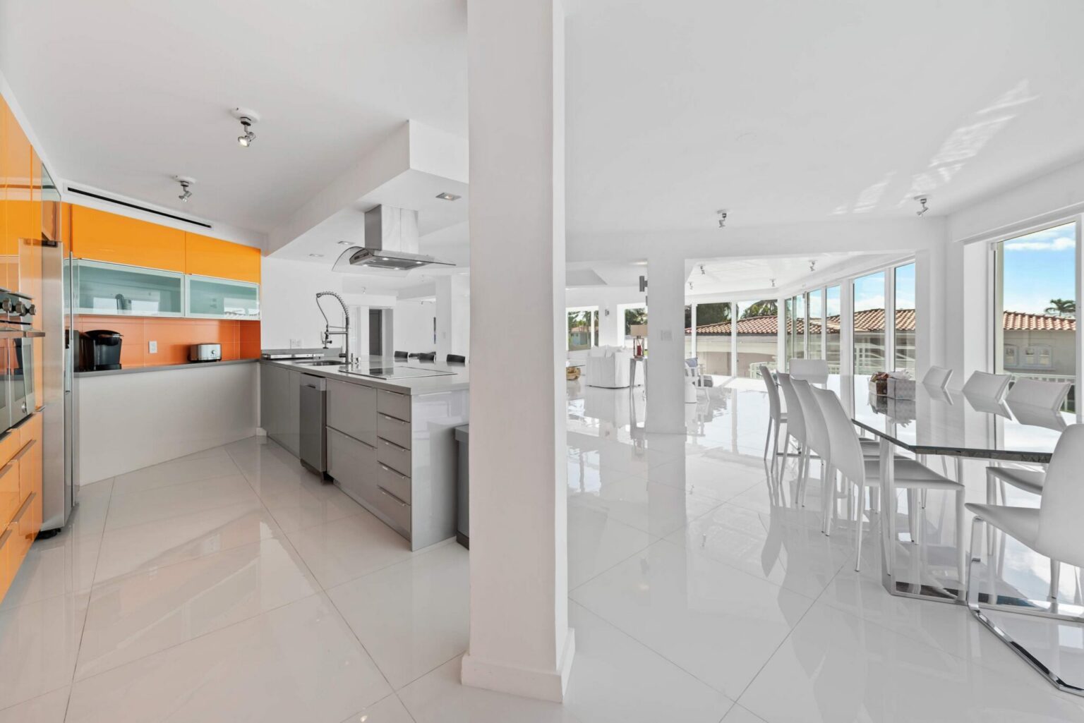 Open-plan kitchen and dining area featuring a chef’s kitchen with stainless steel appliances, custom cabinetry, expansive counter space, a glass dining table, tile flooring, and floor-to-ceiling windows overlooking the outdoor entertaining areas.