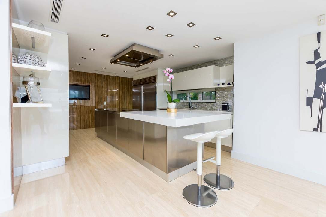 Contemporary kitchen workspace featuring a large waterfall-edge island, stainless steel cabinetry, breakfast seating, custom wood-paneled storage, recessed lighting, built-in appliances, and an open layout connected to the dining and living areas.