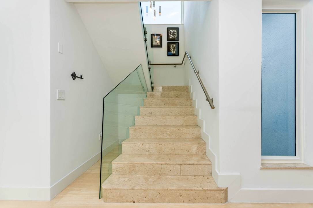 Modern staircase landing featuring natural stone steps, glass railing panels, stainless steel handrails, recessed lighting, contemporary artwork, and a bright open layout connecting multiple levels of the home through a central interior corridor.