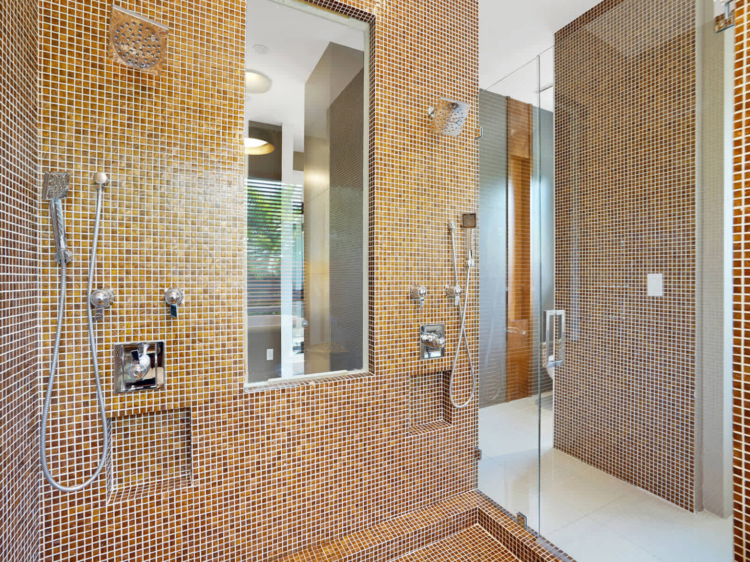 Walk-in shower featuring floor-to-ceiling mosaic tile walls, modern rainfall fixtures, and built-in wall mirrors. Natural light and contemporary finishes enhance the spacious design, creating a bright and functional bathroom space.