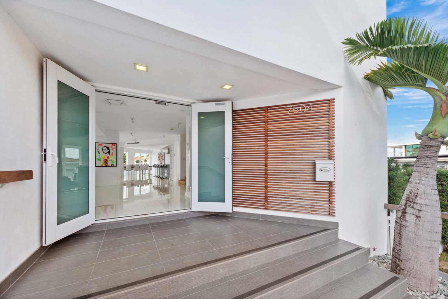 Modern front entrance with oversized glass double doors, tiled entry landing, wood privacy screens, recessed lighting, tropical landscaping, and a covered arrival area that provides direct access to the home's open-concept living spaces.