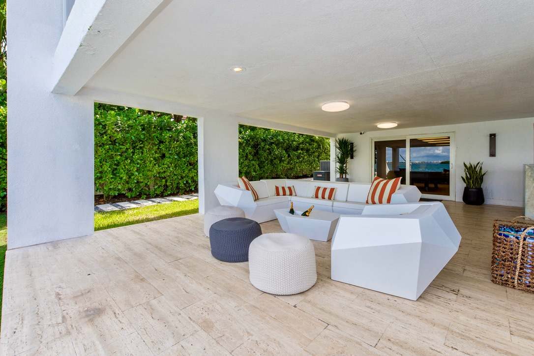 Covered lounge terrace featuring contemporary outdoor seating, cushioned sofas, accent ottomans, wood-look tile flooring, landscaped privacy hedges, recessed lighting, and a spacious open-air layout with direct access to the pool and garden areas.