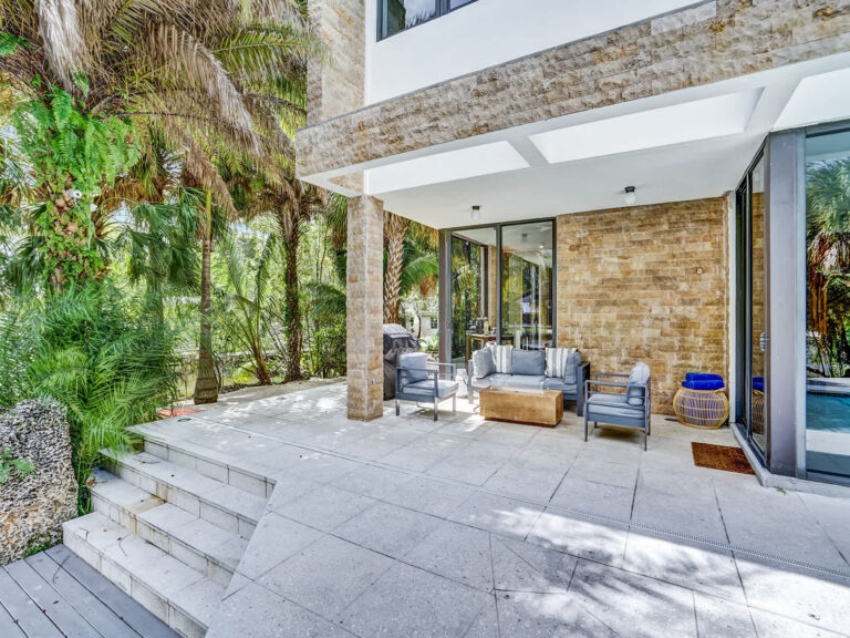 Covered outdoor patio with cushioned lounge seating, stone accent columns, and a spacious tiled terrace. Surrounded by tropical landscaping and mature trees, this shaded gathering area offers seamless indoor-outdoor living.