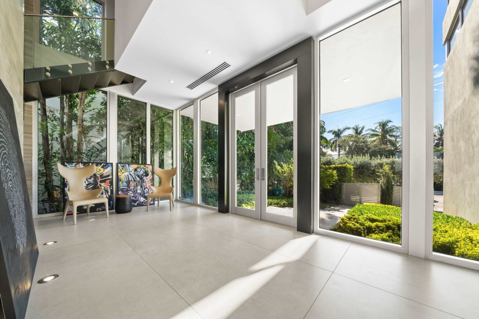 Double-height foyer featuring floor-to-ceiling windows, contemporary architecture, polished tile flooring, and tropical garden views. Natural light fills the space, creating an open entryway with seamless indoor-outdoor connectivity.