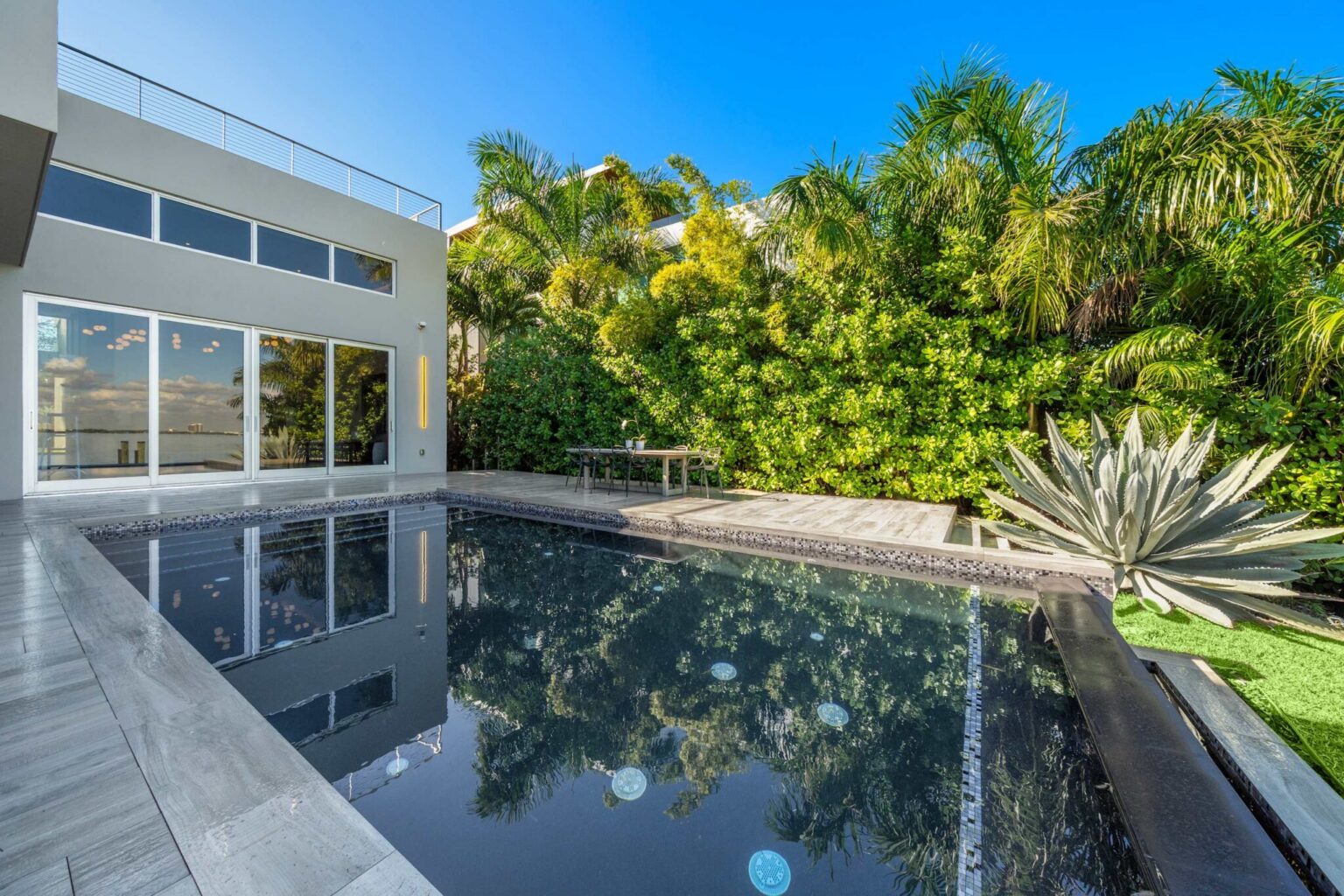 Resort-style swimming pool bordered by tropical landscaping and privacy hedges. Spacious pool deck and modern glass façade create a private outdoor living area for lounging, entertaining, and enjoying the surroundings.