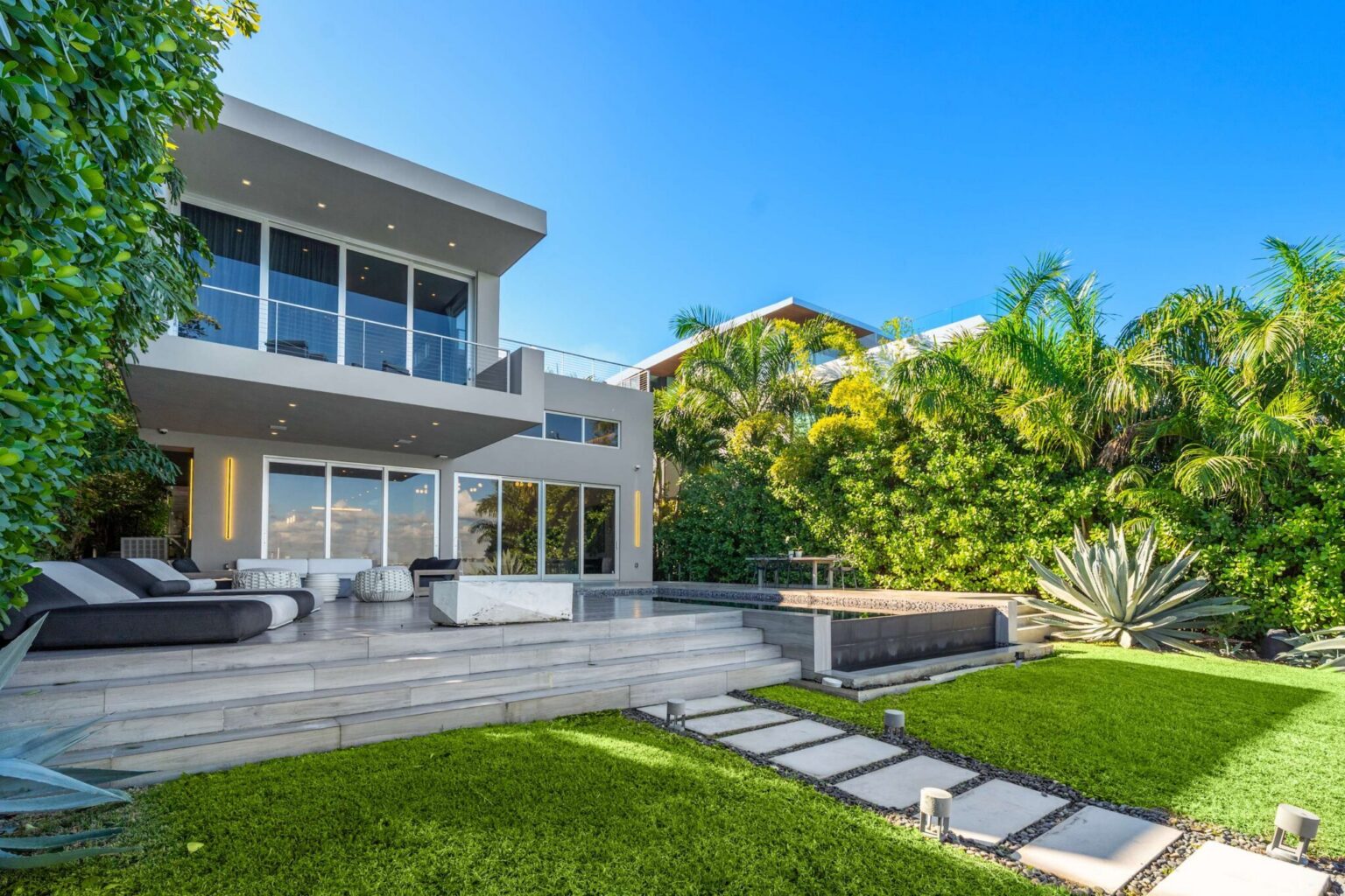 Modern waterfront residence featuring a private pool, expansive sun deck, manicured lawn, and tropical landscaping. Multiple outdoor living areas connect seamlessly to the home's interior through floor-to-ceiling glass doors.