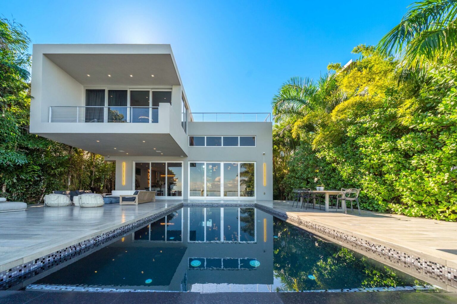Resort-style backyard featuring a private swimming pool, expansive sun deck, tropical landscaping, and contemporary outdoor living spaces. Floor-to-ceiling glass doors connect the outdoor area to the home's modern interiors, creating seamless indoor
