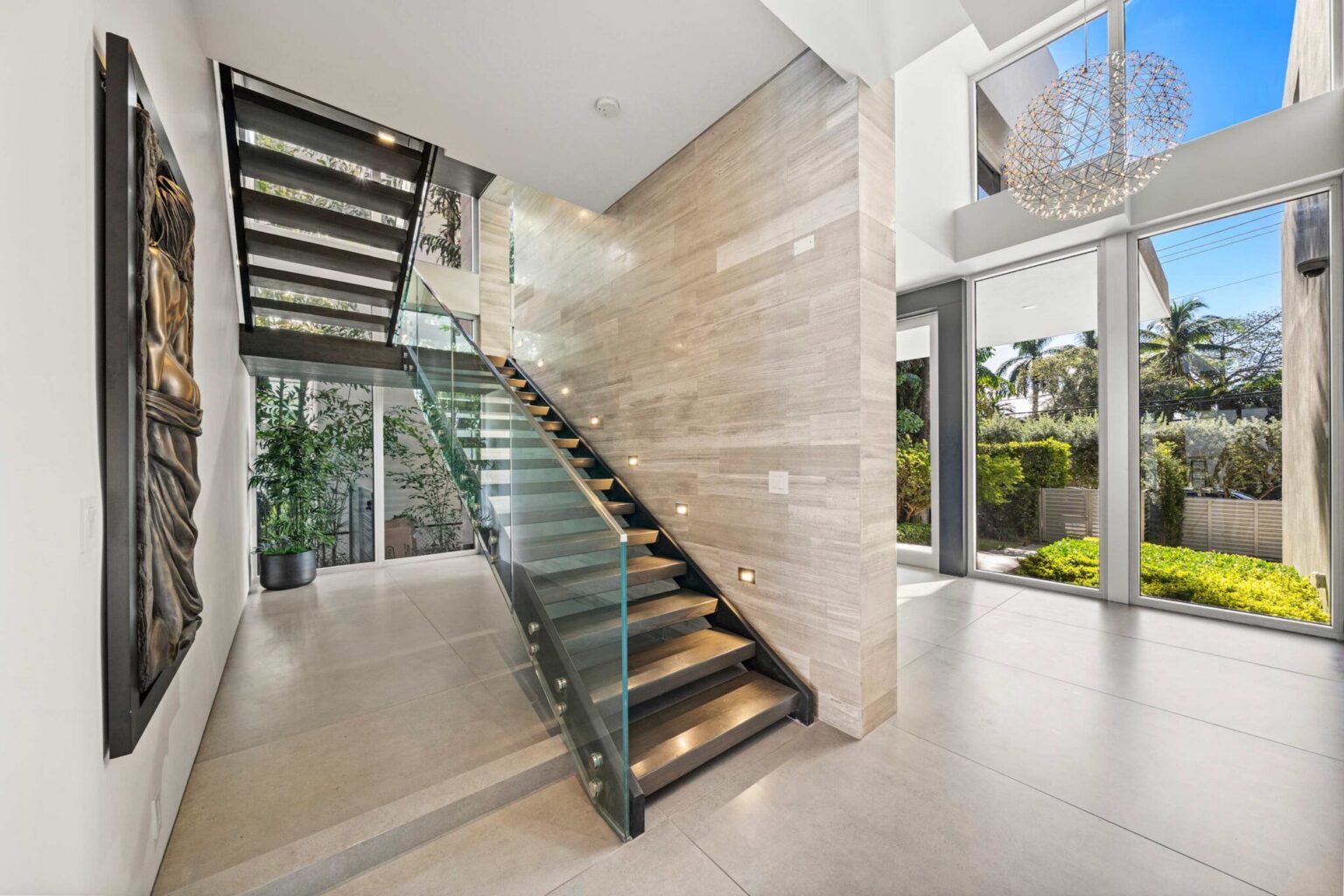 Architectural foyer featuring a floating staircase, glass railings, double-height ceilings, and floor-to-ceiling windows. Natural light, stone accent walls, and contemporary design create a dramatic entryway connecting the home's living spaces.