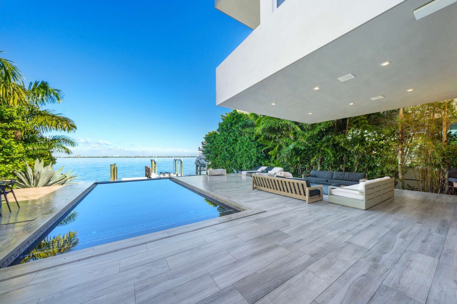 Covered waterfront terrace featuring a private pool, outdoor lounge seating, panoramic bay views, and direct dock access. Expansive deck space, tropical landscaping, and seamless indoor-outdoor living create a spacious waterfront entertaining area.