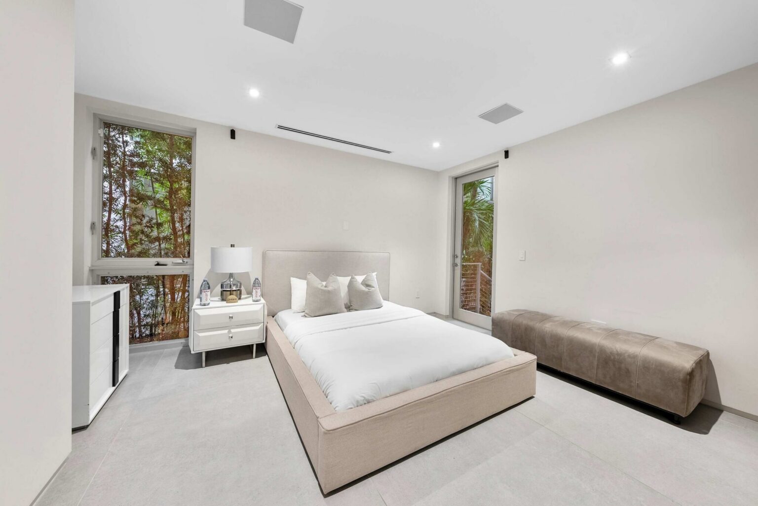 Guest bedroom featuring a queen-size bed, ensuite bathroom access, tile flooring, and contemporary furnishings. Large windows provide natural light and tropical views, while the spacious layout offers comfort and privacy for guests.