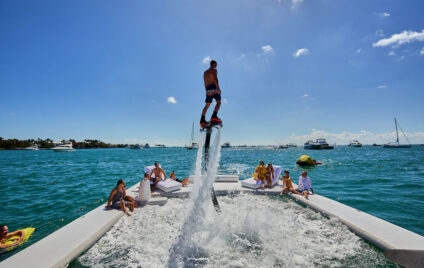 Guests enjoying a luxury yacht party in Miami with a flyboard rider soaring above the water on a sunny day.