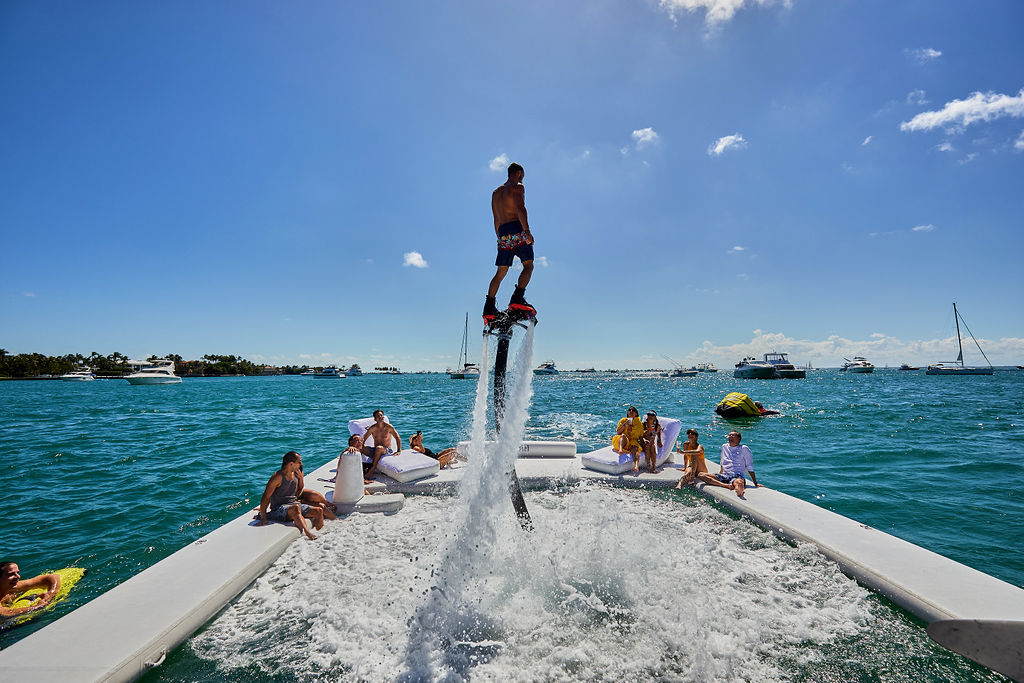 Guests enjoying a luxury yacht party in Miami with a flyboard rider soaring above the water on a sunny day.