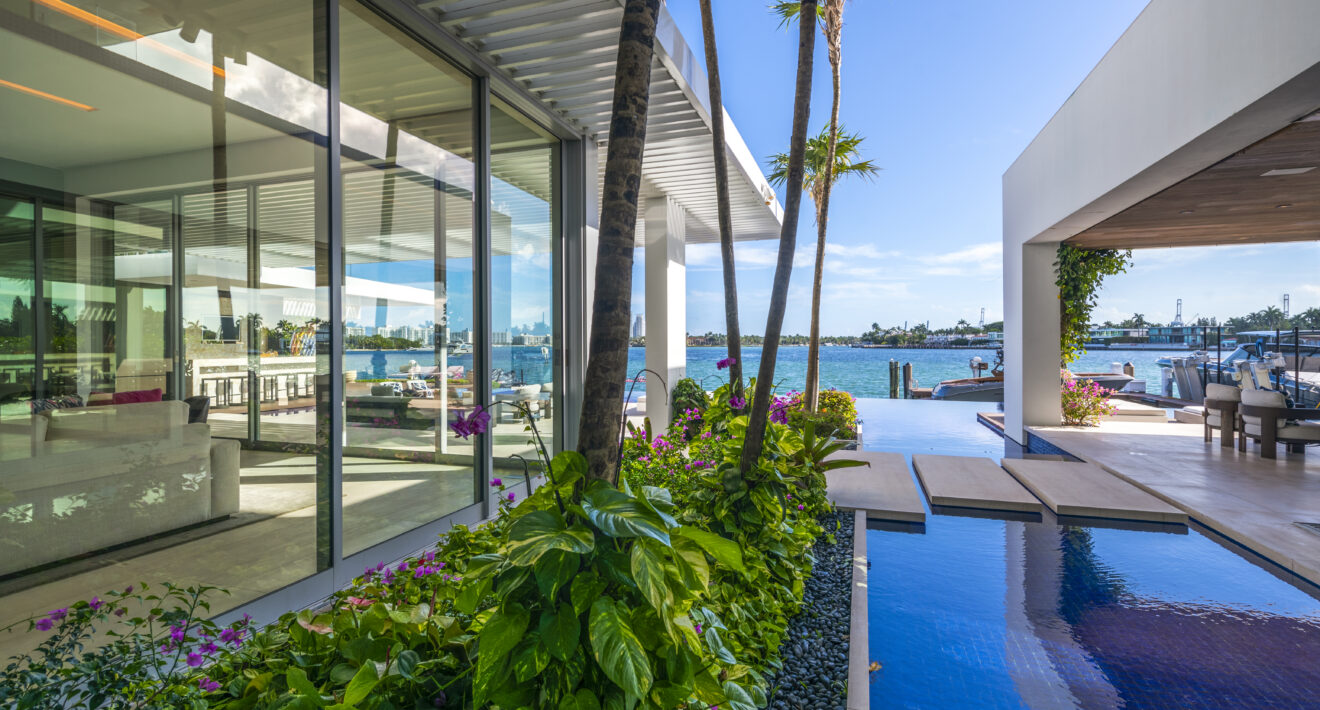 Luxury waterfront Miami villa with floor-to-ceiling glass, tropical landscaping, reflecting pool, and private dock overlooking Biscayne Bay.