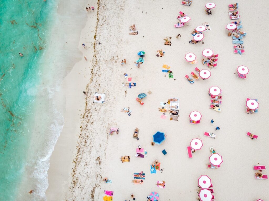 Aerial view of a Miami beach with turquoise water, white sand, colorful umbrellas, lounge chairs, and beachgoers relaxing along the shoreline.