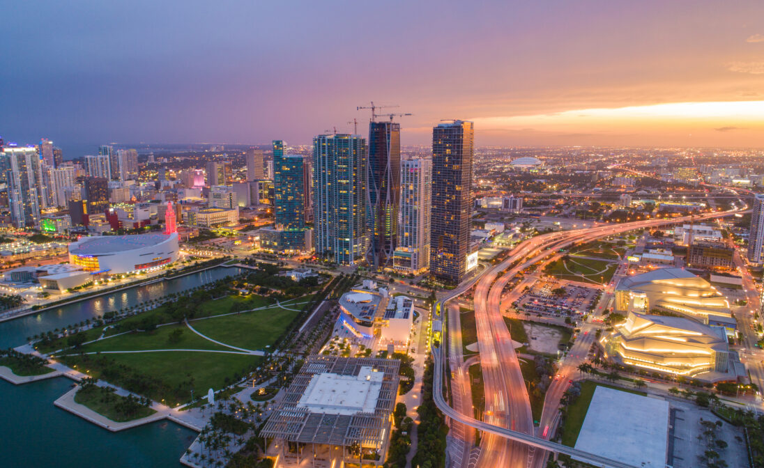 Aerial view of Downtown Miami at sunset with high-rise buildings, waterfront parks, and illuminated city streets.