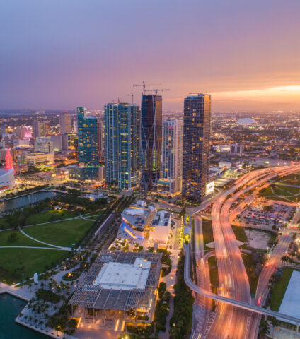 Aerial view of Downtown Miami at sunset with high-rise buildings, waterfront parks, and illuminated city streets.